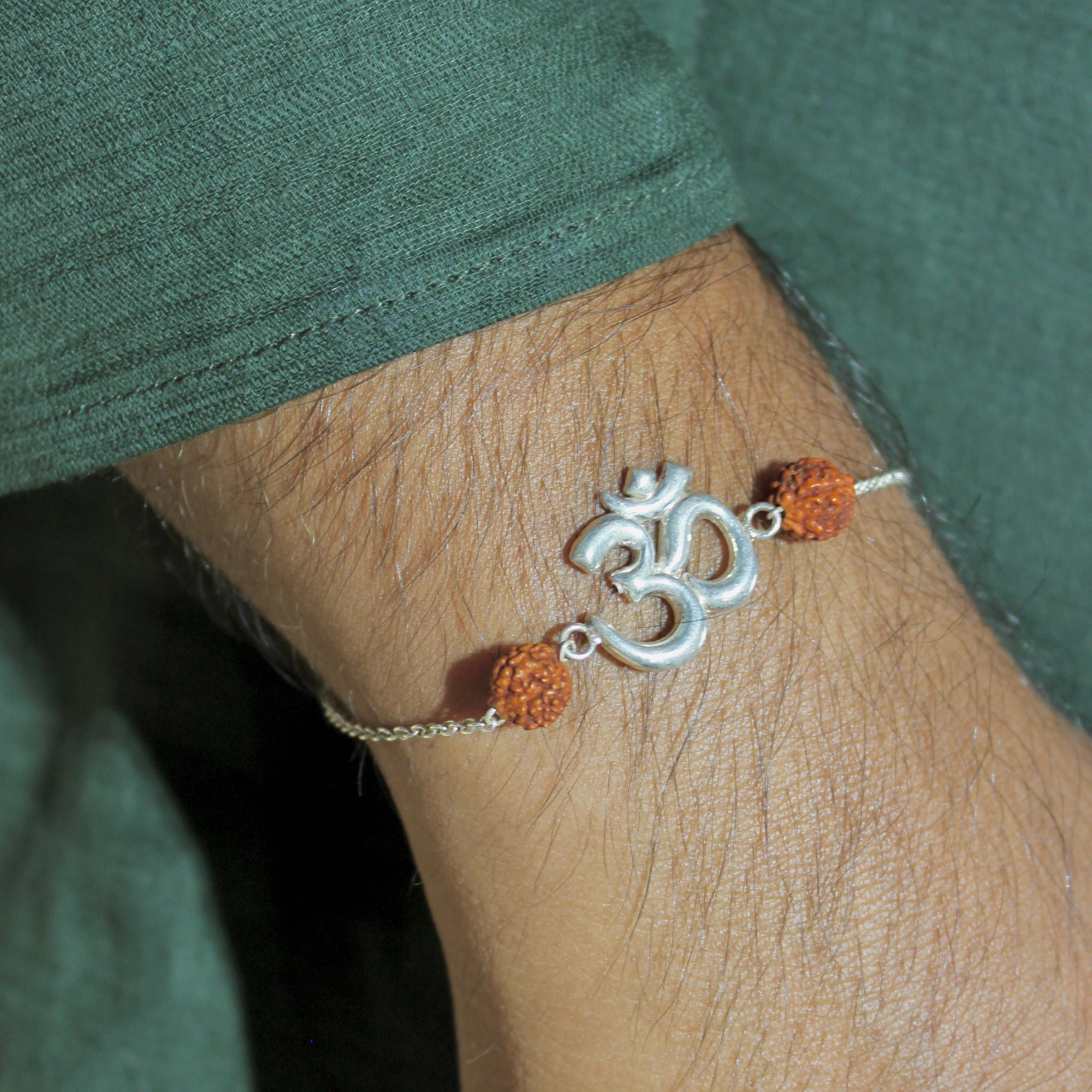 A close-up of a person's wrist wearing a delicate silver-toned chain bracelet with a central Om symbol charm and two small orange beads, set against a dark green shirt.