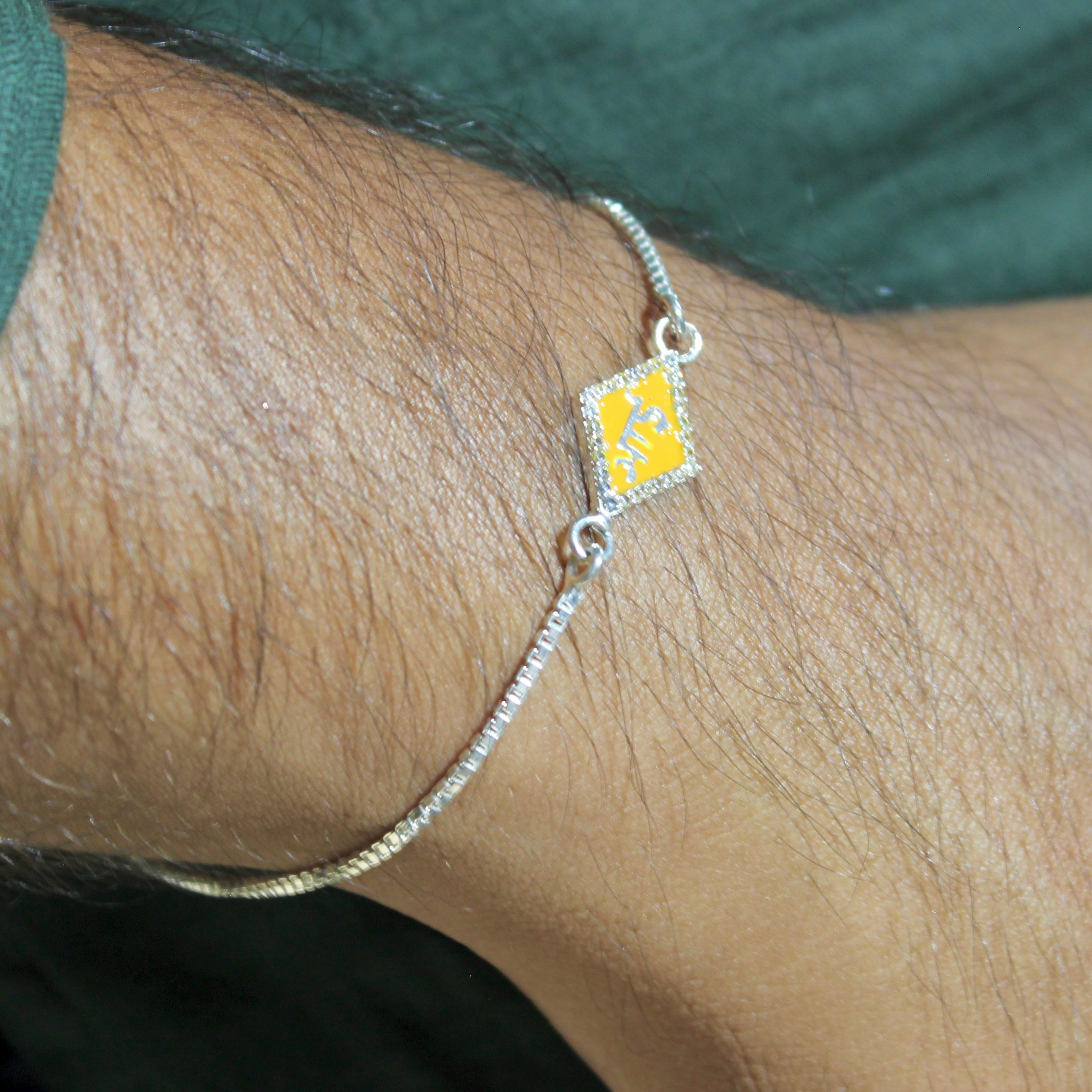 A silver-toned chain bracelet with a diamond-shaped pendant featuring a gold-toned orange center and the initials 'JTS' in gold, resting on a green leaf with a wooden box in the background.