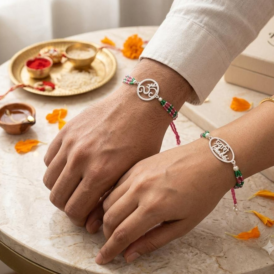Two silver Om bracelets with pink and green beaded straps are shown on a person's wrists, resting on a marble table with traditional Indian ceremonial props and a Parva gift box.