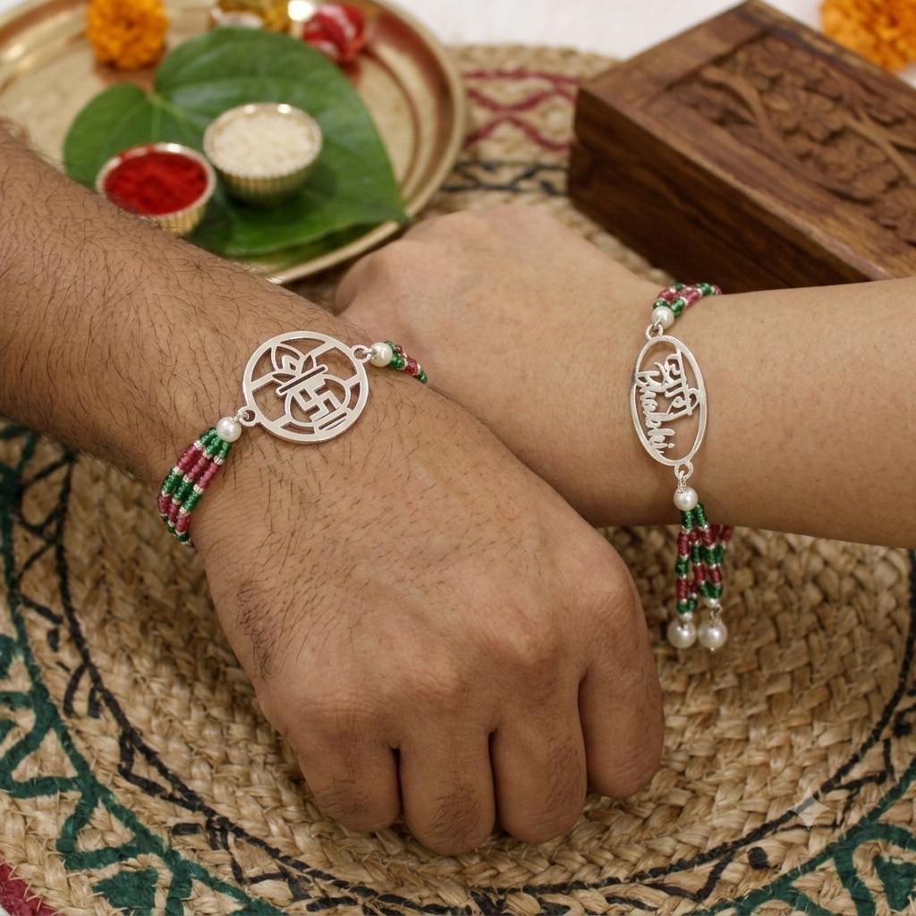 Two silver-toned bracelets with circular and oval motifs, decorated with red and green beaded strands and pearl-like drops, shown worn on two wrists. The wrists rest on a woven mat with decorative traditional props in the background.