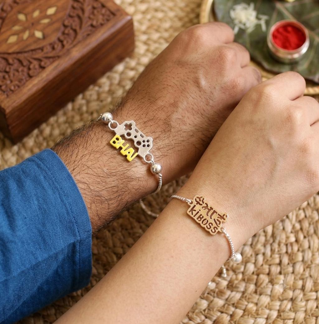 Two hands resting on a woven mat, each wearing a silver bracelet with a gold-toned nameplate. One bracelet reads 'BHAI' and the other reads 'KHOSS' in Devanagari script, with a small pearl bead accent.