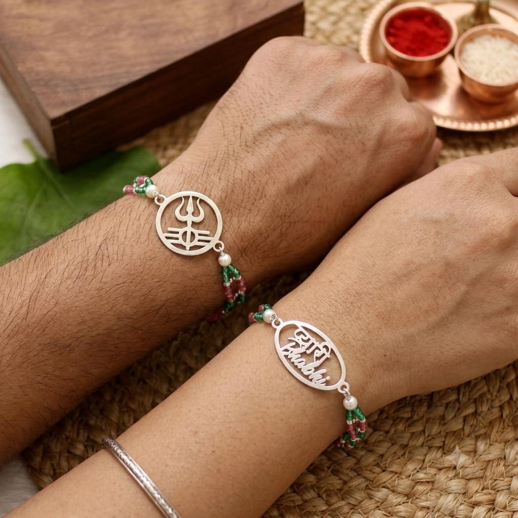 Two silver-toned personalized name and mantra bracelets worn on a person's wrists, styled with green and red beads on a woven mat with traditional decorative elements in the background.