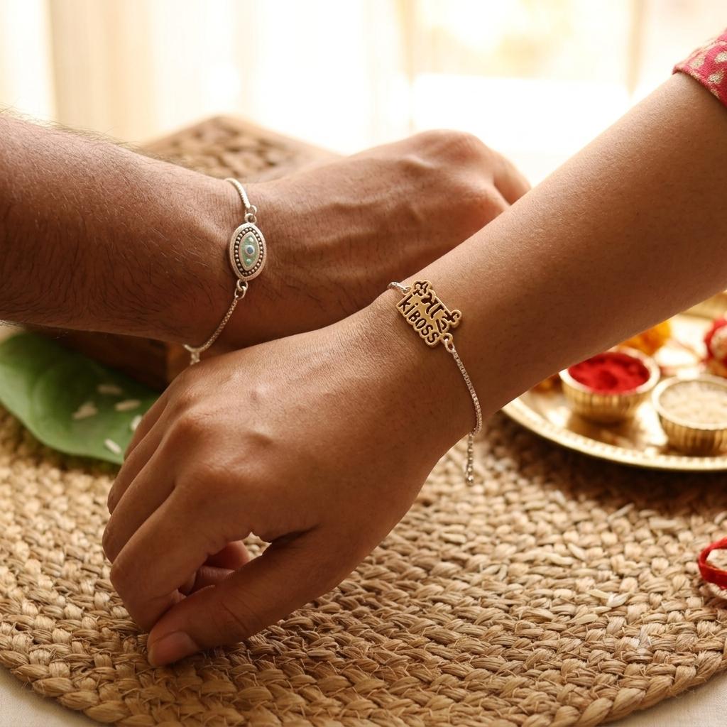 Two people wearing delicate silver bracelets, one with an oval silver charm and the other with a small gold-toned charm engraved with 'Happy Birthday', resting their hands on a woven straw mat.
