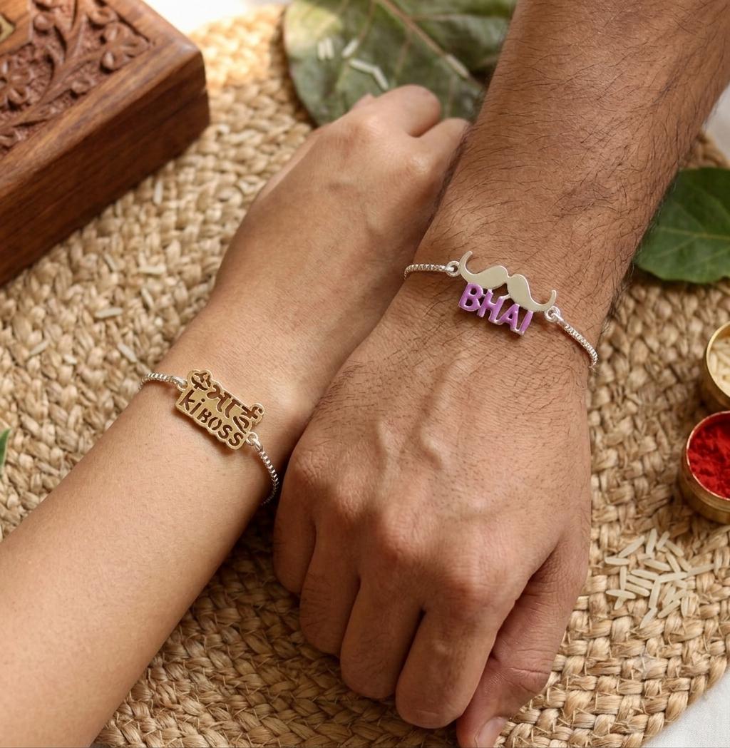 Two silver-toned friendship bracelets on hands, one engraved with 'Bhai Bhaiya KidoS' and the other with a camel and 'Bhai' in purple letters, shown on a woven mat with decorative props.