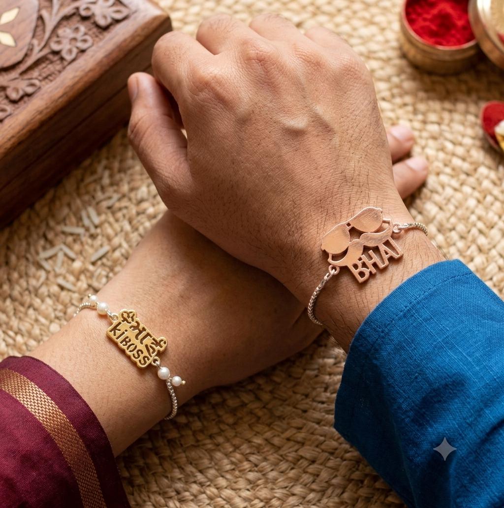 Two hands resting together, each wearing a delicate bracelet. One bracelet is gold with a 'Kumkum Kiss' nameplate and white pearls, and the other is silver with a 'Bhai' elephant charm.